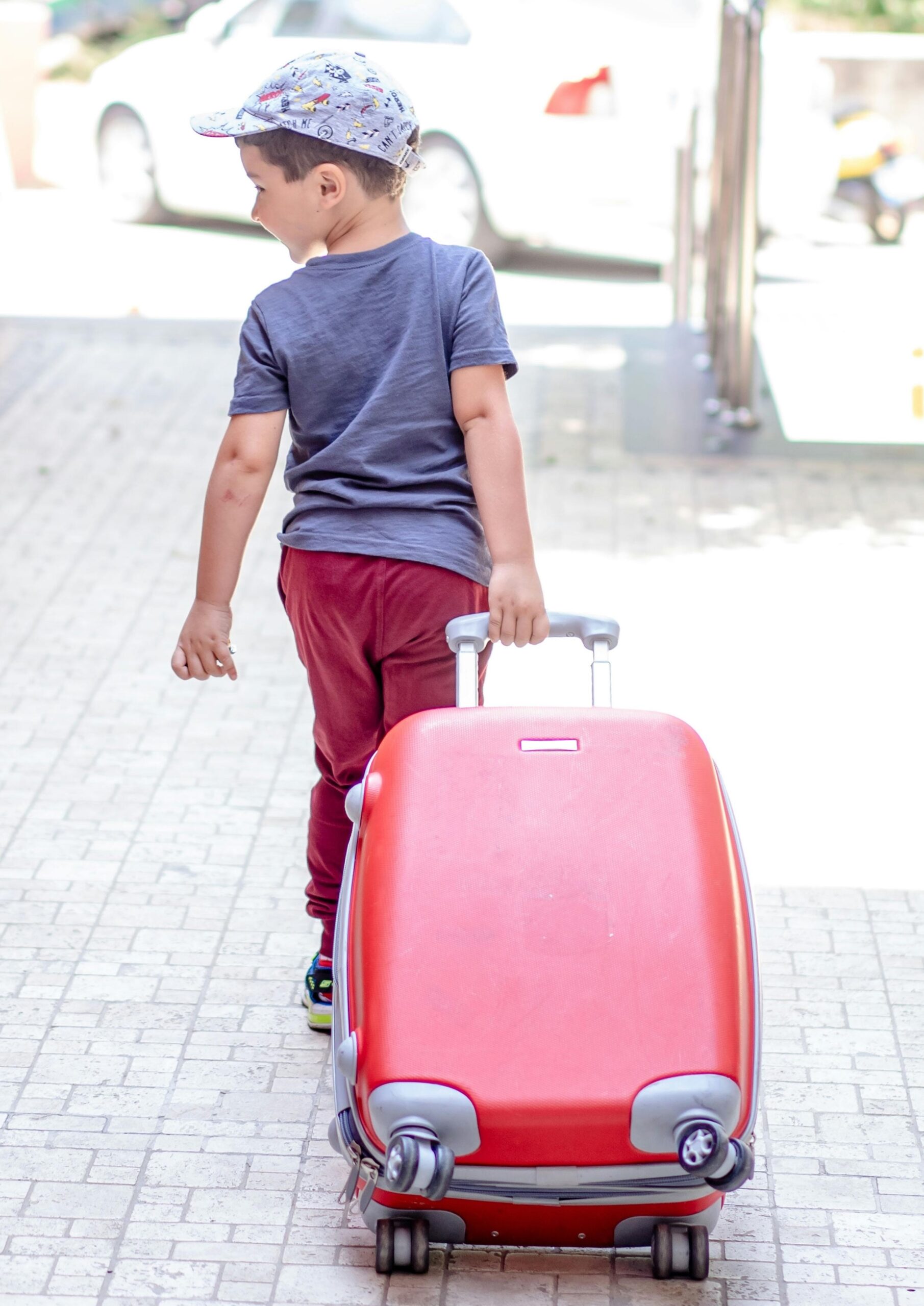 Boy with ball hat walking and pulling a red suitcase.