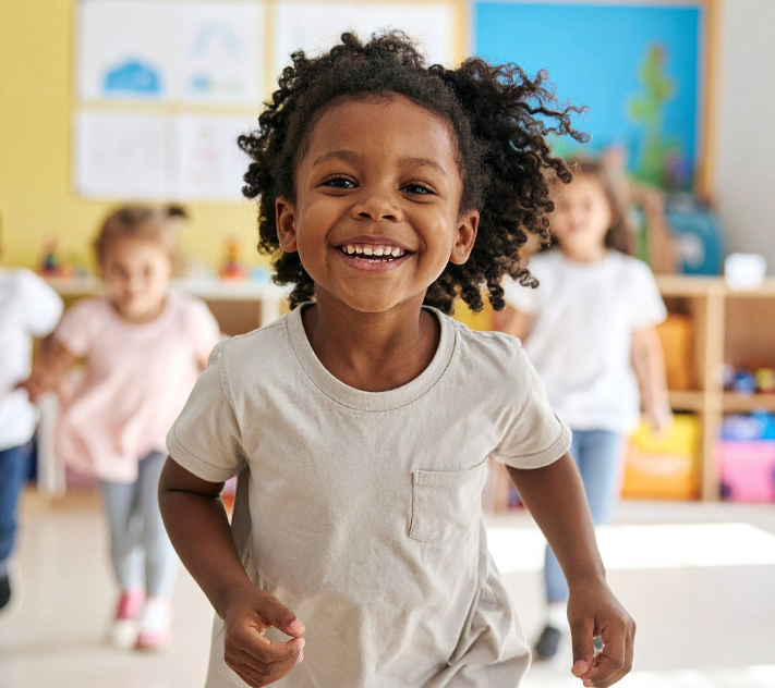 Criança pequena em idade pré-escolar correndo e sorrindo com outras crianças ao fundo.