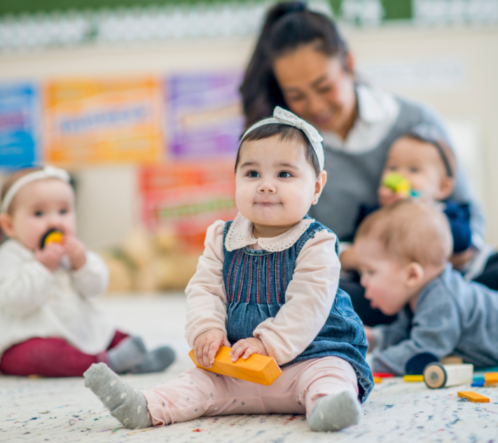 ブロックを手に持った幼い女の子が、絨毯の上に座って微笑んでいる。